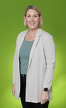 Kerstin Schramm Porträt einer Frau mit kurzen blonden Haaren, die in einem grauen Blazer und einem grünen Oberteil gekleidet ist. Sie lächelt freundlich vor einem leuchtend grünen Hintergrund.