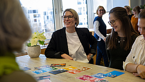 Eine lachende Frau mit Brille sitzt an einem Tisch und spricht mit anderen Frauen. Auf dem Tisch liegen bunte Karten. Im Hintergrund sind mehrere Personen zu sehen, die an einer Besprechung teilnehmen. Die Atmosphäre ist freundlich und entspannt.