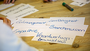 Auf einem Tisch liegen verschiedene Zettel mit handgeschriebenen Begriffen: Zeitmanagement, Lernfähigkeit, Empathie, Branchenwissen und Verantwortungsbewusstsein. Ein Teil einer Hand ist sichtbar, die auf einen Zettel deutet.