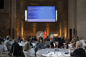 Eine Frau steht auf einer Bühne und spricht zu einem Publikum. Der Titel auf der Leinwand lautet "Nationaler Preis 2025 - Bildung für nachhaltige Entwicklung". Tische sind mit Gästen eingedeckt, während die Frau eine Präsentation hält.
